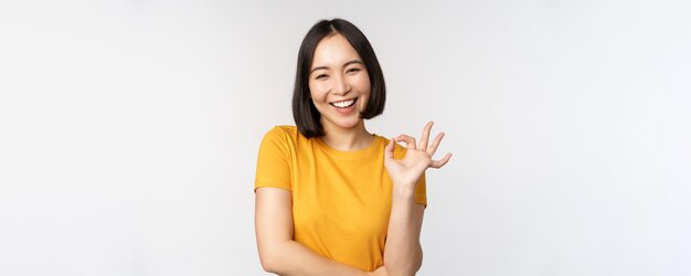 Hermosa mujer joven que muestra el signo de estar bien sonriendo complacida recomendando algo aprobado como producto de pie en una camiseta amarilla sobre fondo blanco