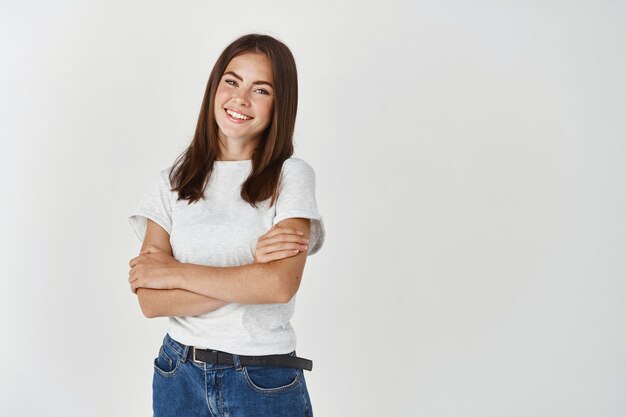 Hermosa mujer joven que parece feliz y despreocupada, de pie con los brazos cruzados sobre el pecho sobre la pared blanca.