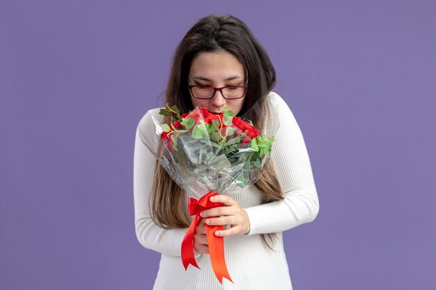 Hermosa mujer joven en ropa casual sosteniendo un ramo de rosas rojas feliz y alegre sonriendo concepto del día de San Valentín de pie sobre la pared púrpura