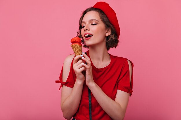 Hermosa mujer joven en ropa roja comiendo helado. Modelo femenino francés refinado posando con postre.