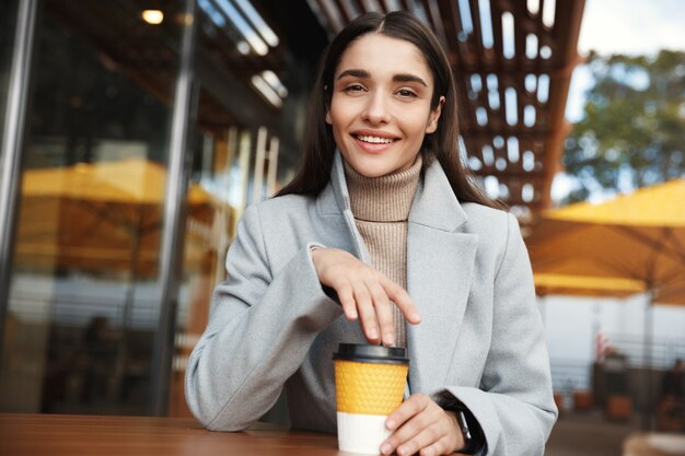 Hermosa mujer joven sentada en abrigo y reloj inteligente esperando en un café.