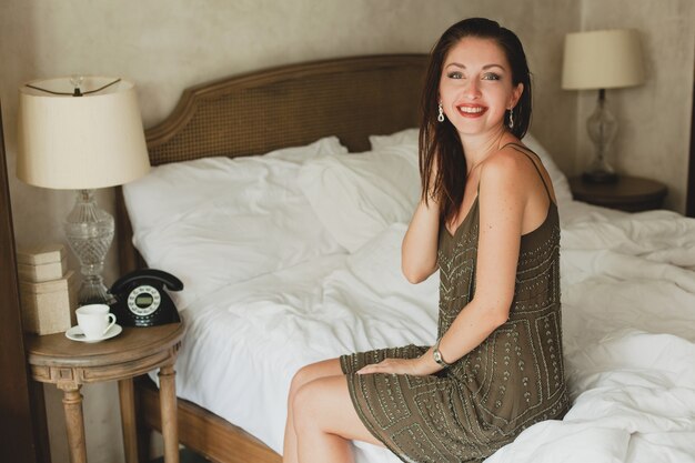 Hermosa mujer joven sentada en la cama en la habitación del hotel, elegante vestido de noche, coqueto, sexy, traje de moda, sábanas blancas