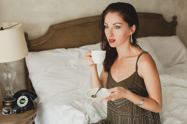 Hermosa mujer joven sentada en la cama en el hotel, vestido elegante, humor sensual, tomando café, sosteniendo la taza