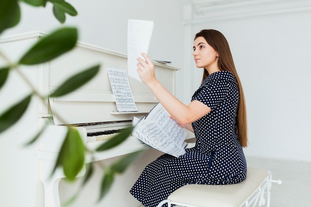 Hermosa mujer joven sentada cerca del piano mirando hoja musical