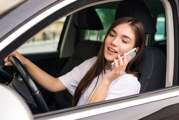 Hermosa mujer joven sentada en el coche con ordenador portátil y hablando por teléfono.