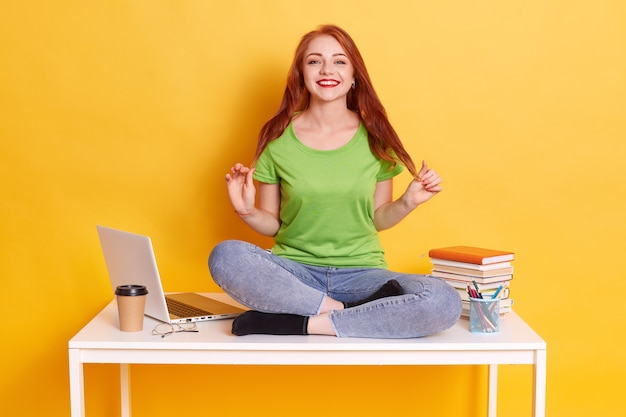 Hermosa mujer joven sentada en la mesa con las piernas cruzadas cerca de la parte superior del regazo blanco, vestidos femeninos atractivos, camiseta casual y jeans, mirando sonriendo a la cámara