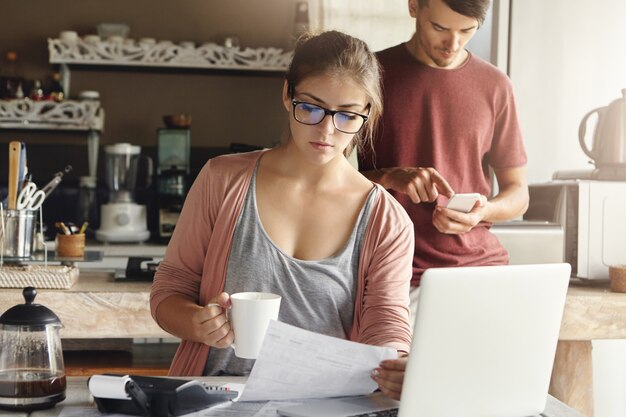 Hermosa mujer joven seria en vasos rectangulares tomando café y estudiando bill en su mano, sentada en el interior de la cocina moderna frente a la computadora portátil abierta