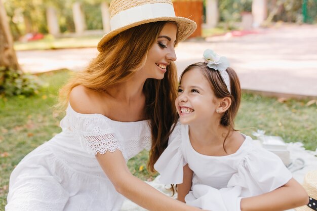 Hermosa mujer joven con sombrero de moda con cinta blanca va a besar a su hija en la frente. Riendo a chica morena con cinta divirtiéndose con mamá pasando el fin de semana en el parque.