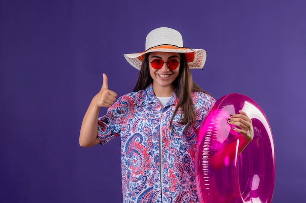 Foto gratuita hermosa mujer joven con sombrero de verano y gafas de sol rojas con anillo inflable con cara feliz sonriendo alegremente mostrando los pulgares sobre la pared púrpura