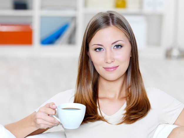 Hermosa mujer joven sonriente con una taza de café en el sofá - en el interior