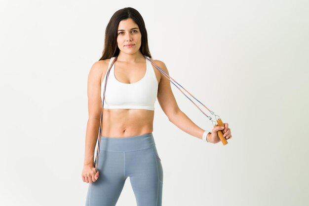 Foto gratuita hermosa mujer joven sosteniendo una cuerda de saltar para hacer ejercicio cardiovascular. atleta en forma en ropa deportiva terminando un entrenamiento hiit frente a un fondo blanco