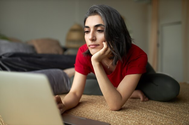 Hermosa mujer joven de stylsih con cabello grisáceo y piercing facial sentada en una alfombra frente a una computadora portátil abierta, navegando por internet usando una conexión inalámbrica, viendo un tutorial de yoga en línea.