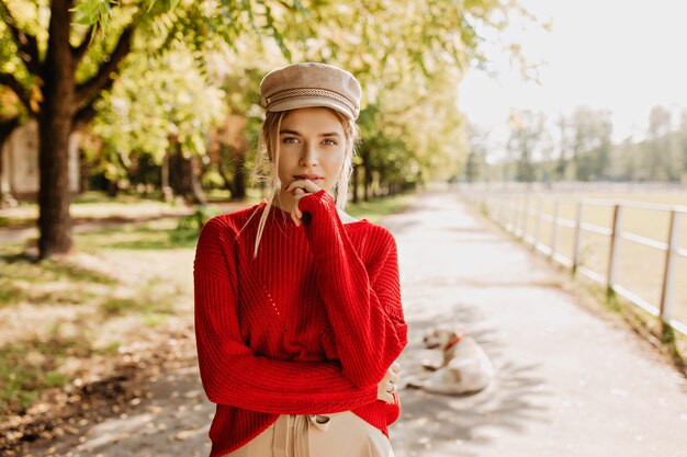 Hermosa mujer joven en suéter rojo y un bonito sombrero de moda mirando pensativo en el parque de otoño. Rubia atractiva en ropa elegante posando al aire libre.