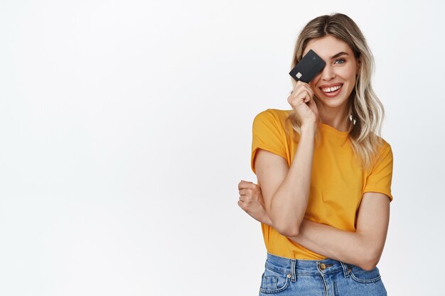 Hermosa mujer joven con tarjeta de crédito en la cara y sonriendo, concepto de compras y pago sin contacto, de pie en blanco.