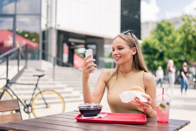 Hermosa mujer joven con teléfono móvil comiendo sabrosa hamburguesa en la cafetería al aire libre