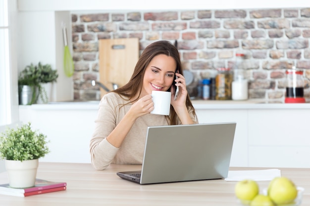 Hermosa mujer joven con teléfono móvil y una computadora portátil en la cocina.