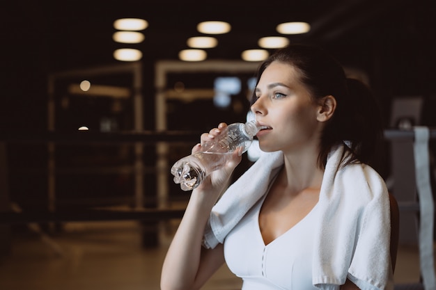 Hermosa mujer joven con una toalla sobre sus hombros bebiendo agua de una botella en el gimnasio