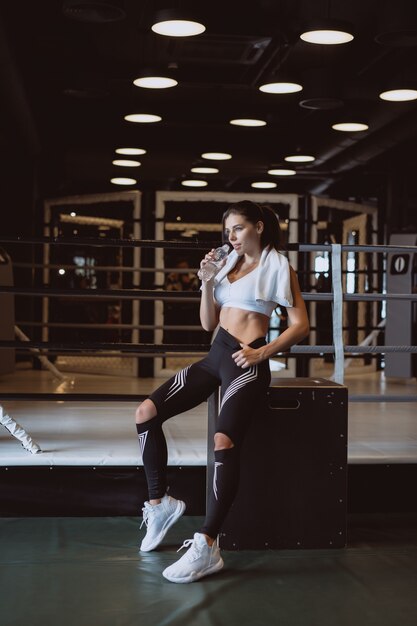 Hermosa mujer joven con una toalla sobre sus hombros bebiendo agua de una botella en el gimnasio