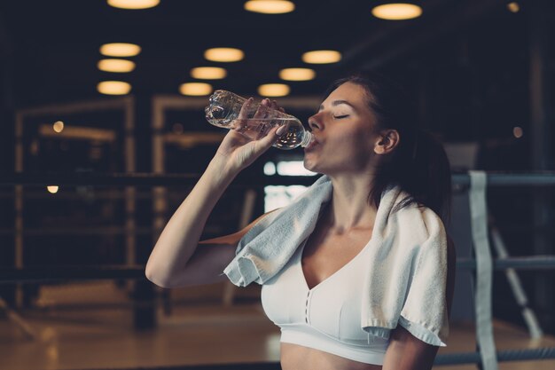 Hermosa mujer joven con una toalla sobre sus hombros bebiendo agua de una botella en el gimnasio