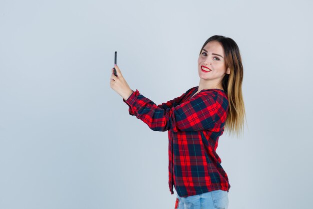 Hermosa mujer joven tomando selfie, mirando en camisa casual y luciendo dichosa. vista frontal.