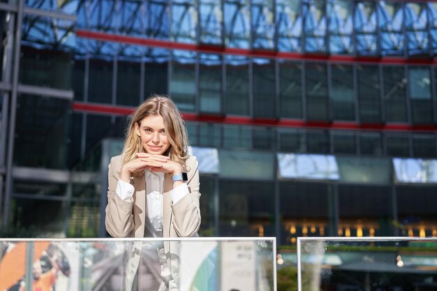 Hermosa mujer joven en traje beige de pie cerca de edificios de oficinas en el centro de la ciudad sonriendo y mirando