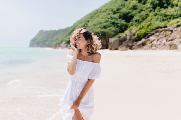 Hermosa mujer joven en traje blanco de pie en la playa tropical. Disparo al aire libre de la atractiva mujer bronceada en vestido posando en la playa de arena.