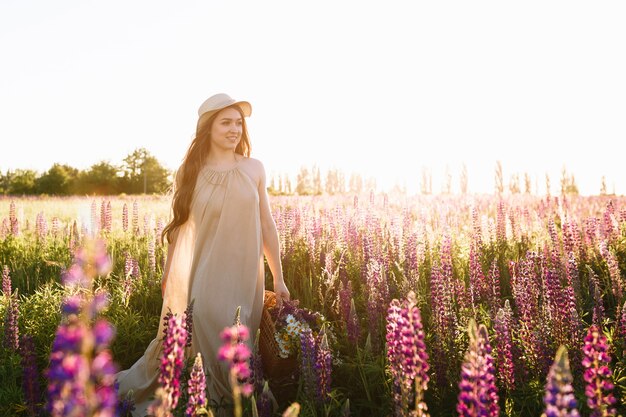 Hermosa mujer joven en vestido blanco y sombrero de paja caminando en campo de flores