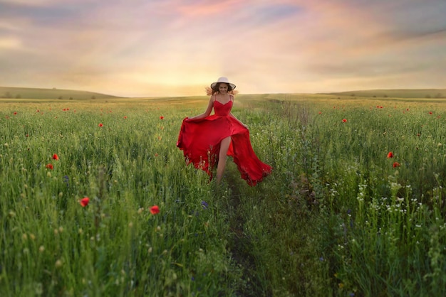 Hermosa mujer joven en vestido rojo y sombrero blanco camina por el campo con amapolas