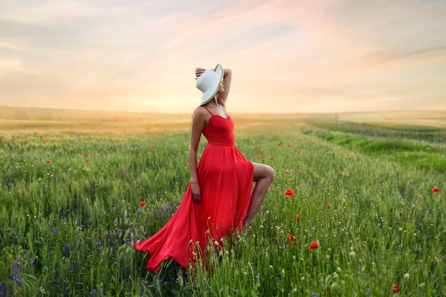 Hermosa mujer joven en vestido rojo y sombrero blanco camina por el campo con amapolas