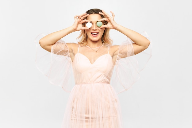 Hermosa mujer joven con vestido rosado endeble posando y sosteniendo macarons en su rostro en lugar de ojos