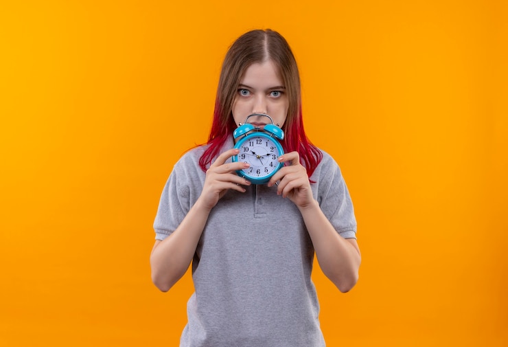 Foto gratuito hermosa mujer joven vistiendo camiseta gris sosteniendo el reloj de alarma en la pared amarilla aislada