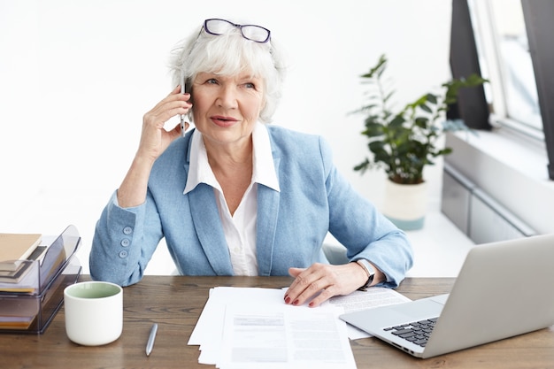 Hermosa mujer madura con cabello gris haciendo llamadas telefónicas en su oficina, elegante empresaria senior en elegante traje hablando por móvil con socio potencial, sentada en el lugar de trabajo con laptop