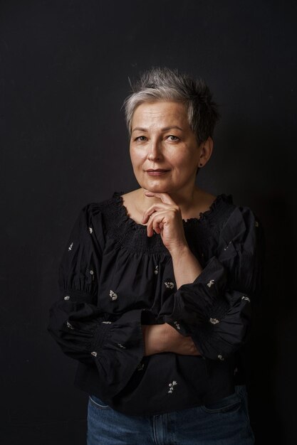 Hermosa mujer madura con cabello gris posando barbilla inclinada sobre su mano aislada sobre fondo negro Mujer elegante gris con belleza corta Envejecida En blanco o plantilla con espacio de texto a la izquierda