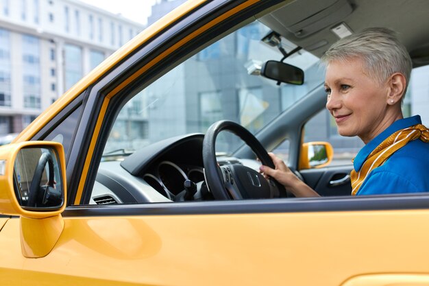 Hermosa mujer madura con cabello pixie conduciendo un auto, disfrutando de calles vacías temprano en la mañana. Atractiva mujer de mediana edad sentado en el asiento del conductor, estacionamiento de automóviles, mirando al espejo retrovisor lateral