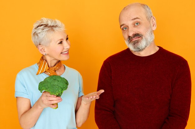 Hermosa mujer madura que cuida con brócoli que ofrece comida saludable a su marido enfermo pensionista que está en una estricta dieta vegetariana. A un anciano disgustado no le gusta comer verduras