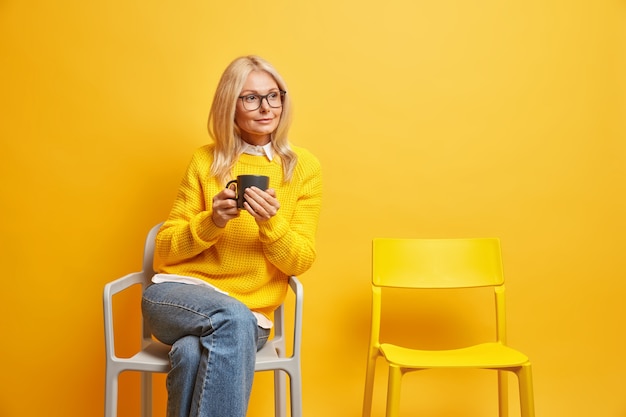 Hermosa mujer mayor de cincuenta años disfruta de tiempo libre para buenos recuerdos, bebidas, té o café, posa en una silla con expresión pensativa y recuerda toda la vida concentrada a un lado. Serenidad en casa