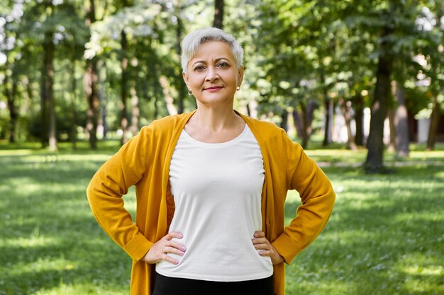 Hermosa mujer mayor de pelo gris en cardigan amarillo y camiseta blanca posando en el parque de verano verde, manteniendo las manos en la cintura, haciendo ejercicios físicos, con sonrisa segura