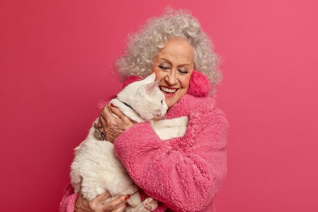 Hermosa mujer mayor de pelo gris lleva un gato blanco en las manos, se preocupa por su propia mascota con amor, se viste de manera informal, aislada sobre una pared rosa, pasa el tiempo libre juntos, se queda en casa durante la cuarentena