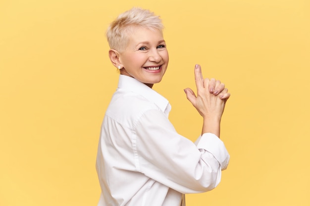 Hermosa mujer de mediana edad alegre con el pelo corto teñido mientras juega con sus nietos haciendo gestos como si sostuviera una pistola, divirtiéndose, posando aislado