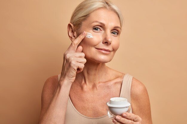 Hermosa mujer de mediana edad aplica crema antienvejecimiento en la cara se somete a tratamientos de belleza se preocupa por la piel