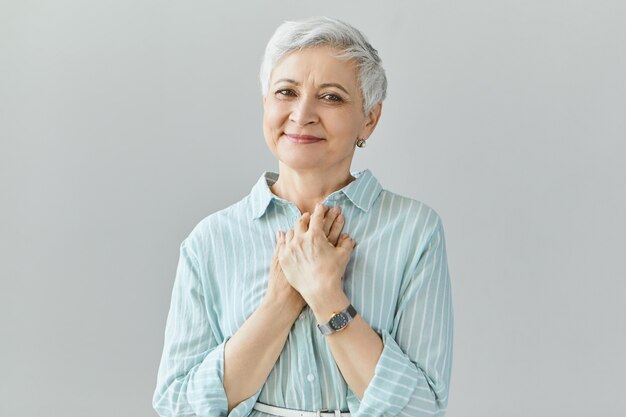 Hermosa mujer de mediana edad de aspecto amistoso con una sonrisa sincera, que expresa gratitud, se siente agradecida, muestra su corazón lleno de amor, manteniendo las manos en el pecho. Sentimientos humanos genuinos positivos