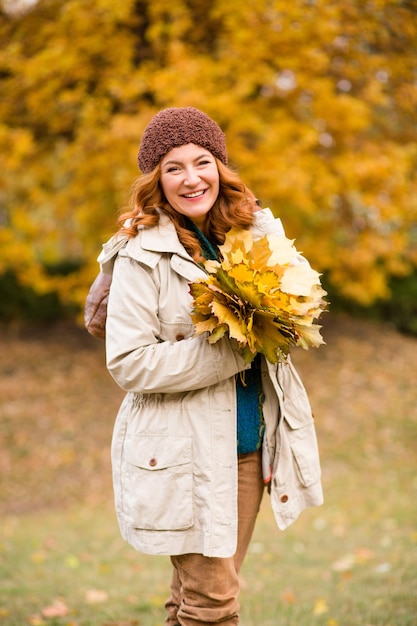 Hermosa mujer de mediana edad caminando en el parque de otoño. Mujer sonriente sosteniendo muchas hojas de arce amarillas y mirando a la cámara.