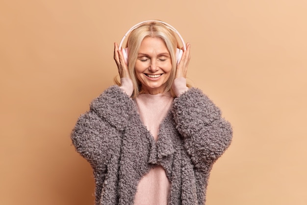 Hermosa mujer de mediana edad feliz escucha música favorita en auriculares mantiene los ojos cerrados y sonríe con satisfacción usa un abrigo cálido pasa el tiempo libre escuchando canciones agradables posa en el interior