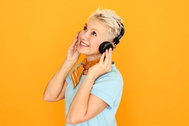 Hermosa mujer de mediana edad con ojos azules y cabello corto posando sobre fondo amarillo mirando hacia arriba con una alegre sonrisa alegre, escuchando música con auriculares inalámbricos.