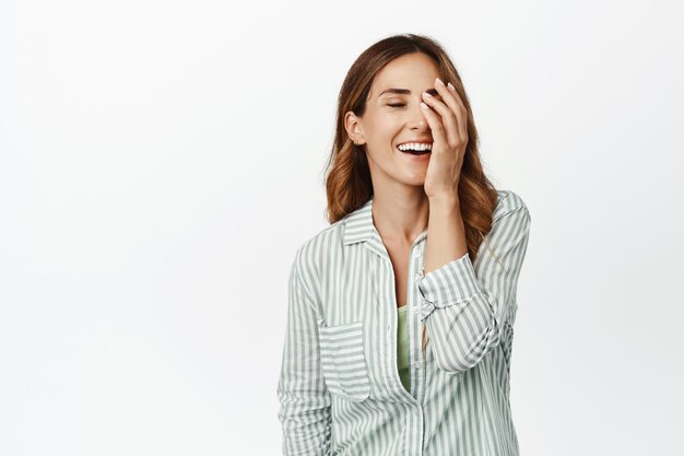 Hermosa mujer de mediana edad con piel perfecta, sin arrugas, sonriendo y riendo, tocándose la cara, parada en blusa contra fondo blanco