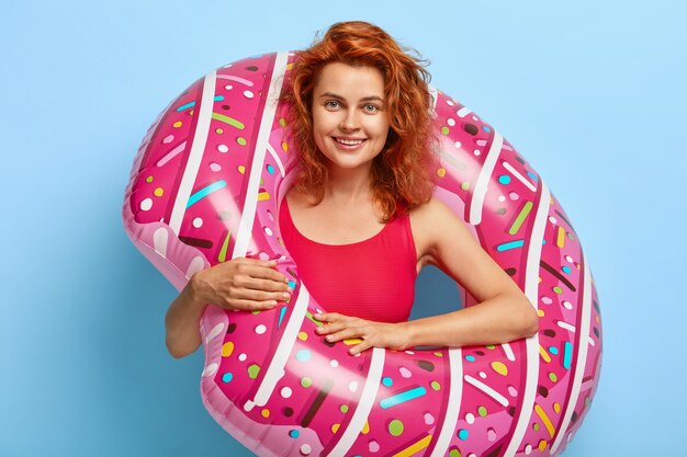Hermosa mujer milenaria con cabello rojo ondulado posando contra la pared azul con donut flotante