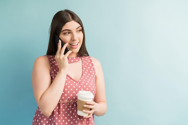 Hermosa mujer milenaria hablando por teléfono inteligente mientras sostiene una bebida contra un fondo liso