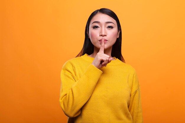 Hermosa mujer misteriosa con suéter amarillo vibrante haciendo gesto de silencio sobre fondo naranja. Persona reservada tocando los labios con el dedo índice indicando silencio y secreto.