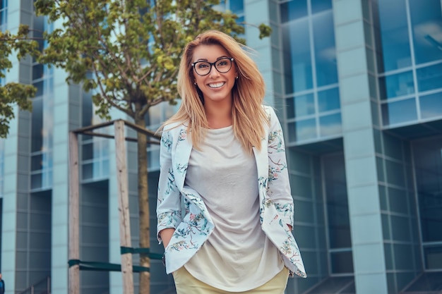 Hermosa mujer de moda sonriente con ropa y gafas elegantes, parada contra un rascacielos.