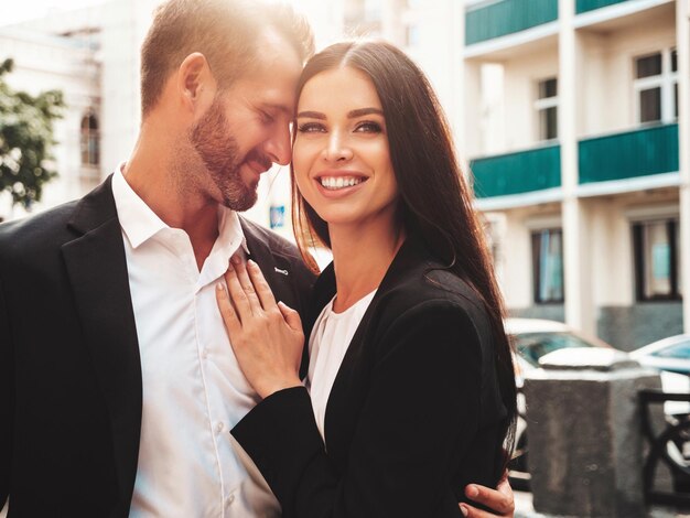 Hermosa mujer de moda y su guapo novio en traje Sexy modelo morena sonriente en vestido de noche negro Pareja de moda posando en la calle al atardecer Hombre brutal y su mujer al aire libre